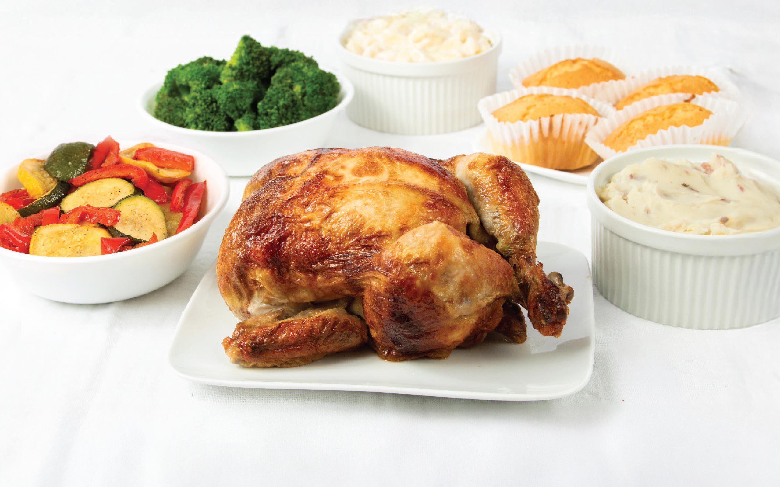 A Rotisserie Chicken Family Meal Deal featuring a whole roasted chicken on a white platter, surrounded by bowls of mashed potatoes, Asiago mac and cheese, roasted vegetables, and steamed broccoli, with a side of six mini cornbread loaves.