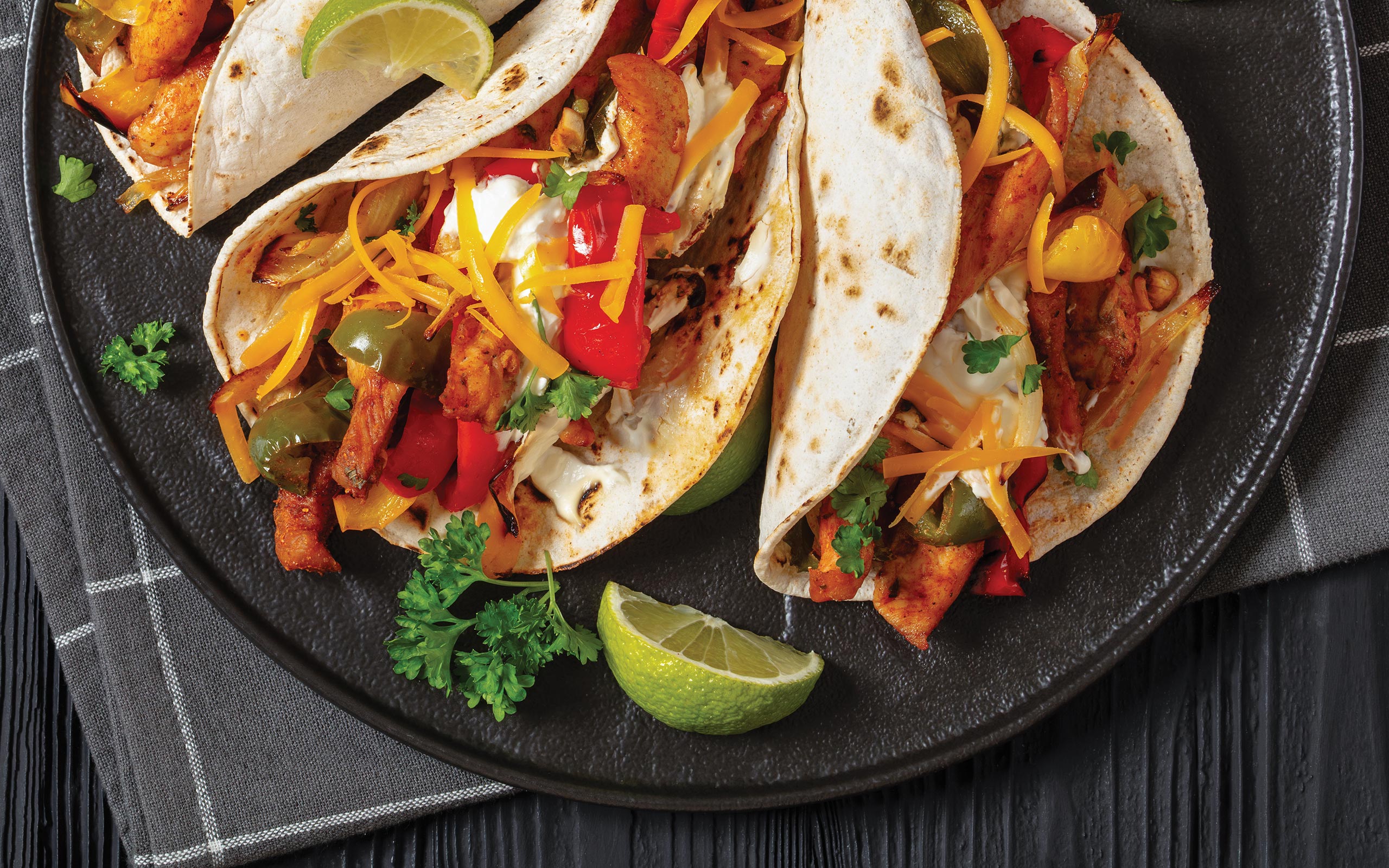 Three chicken fajitas in flour tortillas rest on a dark plate, filled with seasoned chicken, charred red and green bell peppers, and onions. They are topped with shredded cheddar cheese.