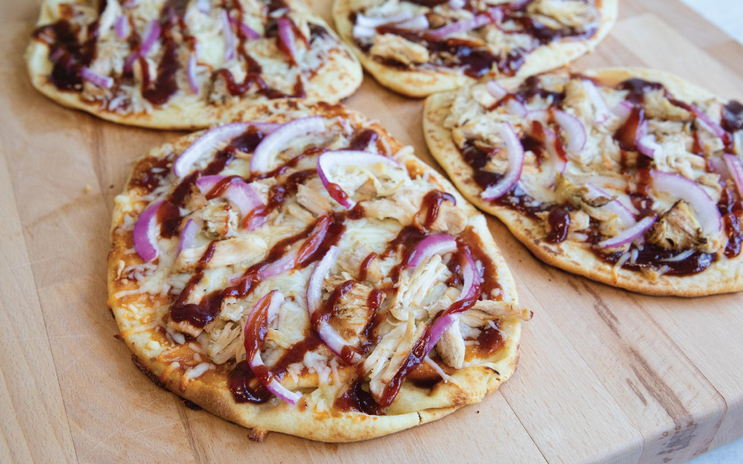 BBQ chicken flatbreads topped with melted cheese, red onion slices, and drizzled barbecue sauce, arranged on a wooden board.
