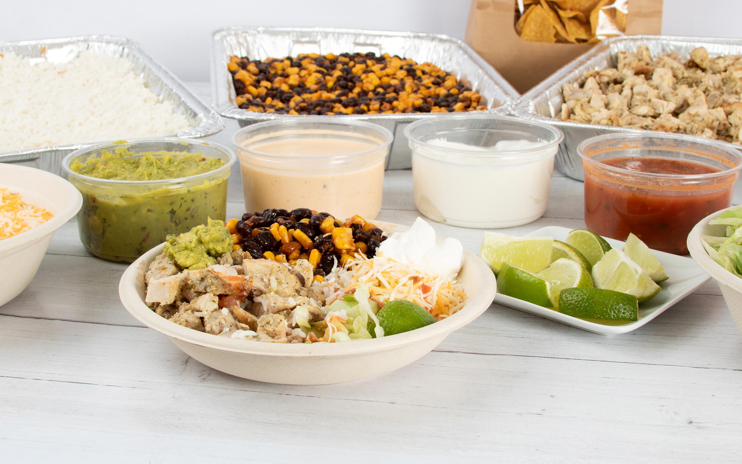 A prepared burrito bowl filled with cilantro lime chicken, rice, black beans, corn, shredded cheese, lettuce, guacamole, and sour cream sits in the foreground. In the background, large aluminum catering trays of rice, black bean and corn blend, and chicken are arranged alongside containers of salsas, guacamole, sour cream, a small plate of lime wedges, and a bag of tortilla chips.