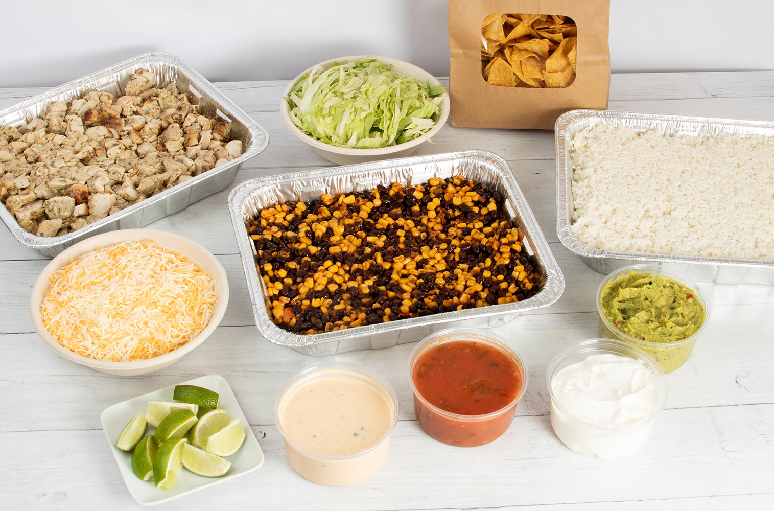 A catered burrito bowl spread featuring large aluminum trays of cilantro lime chicken, jasmine rice, and a southwest black bean and corn blend. Surrounding the main dishes are bowls of shredded cheese, shredded lettuce, a brown paper bag of tortilla chips, fresh lime wedges, and clear containers of guacamole, salsa, sour cream, and a creamy sauce.